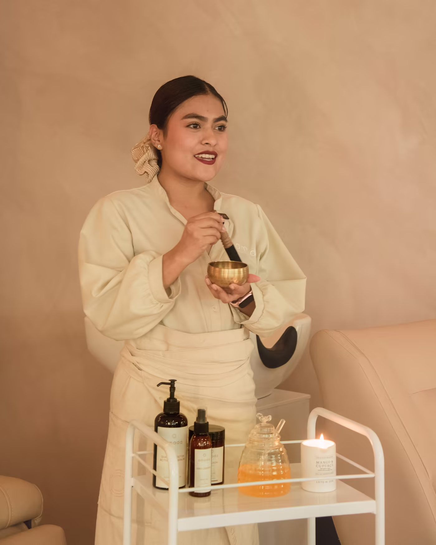 Especialista de Amada con uniforme de lino sostiene un cuenco tibetano junto al carrito del ritual con aceites y una vela