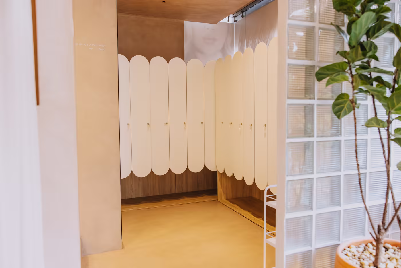 Vestier de Amada con lockers blancos de formas curvas, muro de bloques de vidrio y una planta